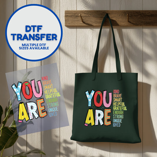 Tote bag with "You Are" design and empowering words, next to a DTF transfer.